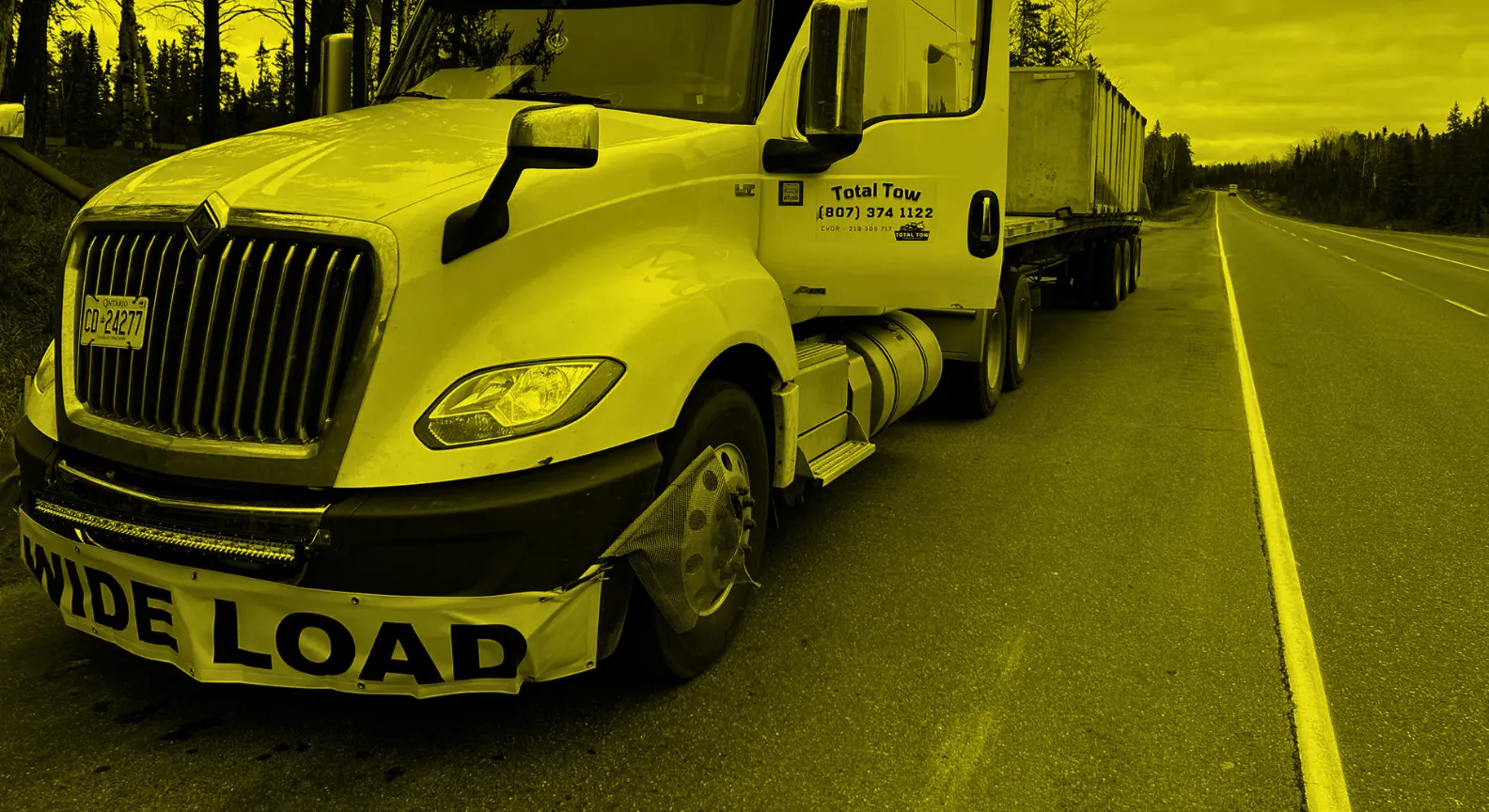 A truck with a WIDE LOAD banner on the front bumper, hauling an oversize trailer on the highway